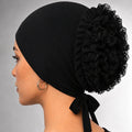 A woman shown in profile wearing a black fabric head covering with a large ruffled pom-pom and side bow, photographed against a neutral gray studio background.