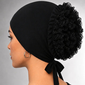 A woman shown in profile wearing a black fabric head covering with a large ruffled pom-pom and side bow, photographed against a neutral gray studio background.