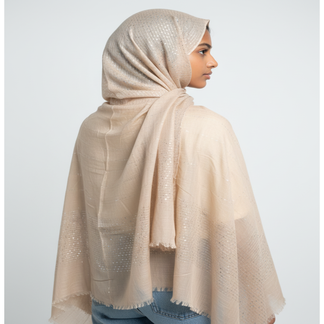 A model shown from the back wearing a light beige shimmering hijab and shawl draped over her shoulders, paired with blue jeans, against a plain light gray background.