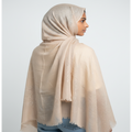 A model shown from the back wearing a light beige shimmering hijab and shawl draped over her shoulders, paired with blue jeans, against a plain light gray background.