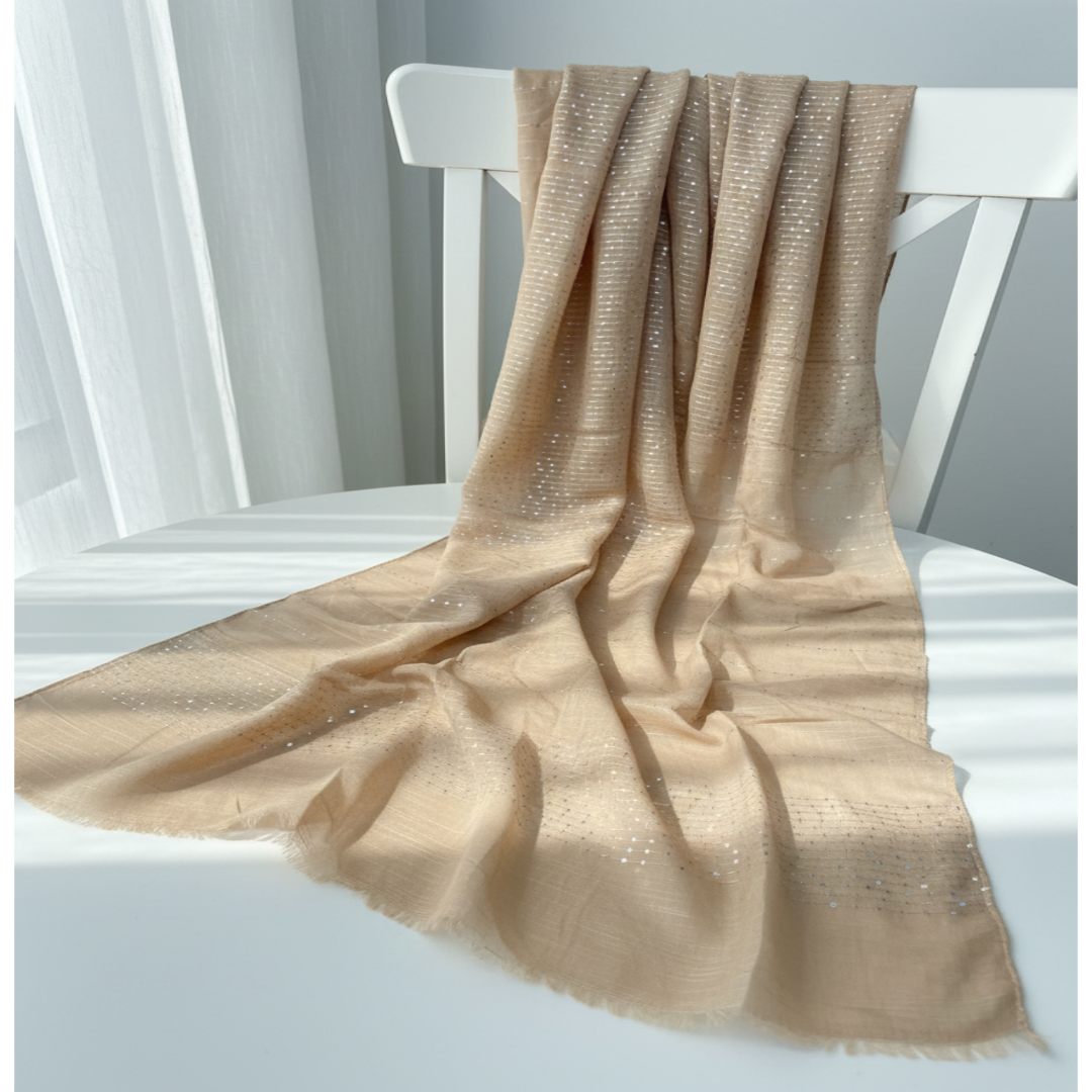 A lightweight beige scarf with subtle silver sequin patterns draped over a white wooden chair in a bright, softly lit room.