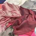 A close-up of draped fabric in multiple colors, with the most prominent piece being a deep burgundy red textile with black fringe, layered over cream, gray, and pink fabrics.