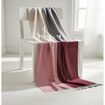 A lightweight woven throw blanket draped over a white chair in a bright, minimalist room. The blanket features a three-color gradient transitioning from cream/off-white at the top, through gray, to dusty rose and deep burgundy, finished with white fringe tassels at the bottom.