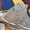 A close-up of a person's hand holding up a crinkled, semi-sheer gray fabric panel with a white ruffled trim, showing its texture and transparency. Other colorful fabric panels are visible in the background.