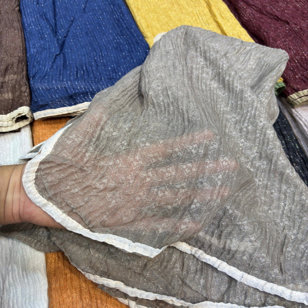 A close-up of a person's hand holding up a crinkled, semi-sheer gray fabric panel with a white ruffled trim, showing its texture and transparency. Other colorful fabric panels are visible in the background.