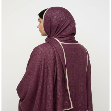 A woman shown from the back wearing a textured plum purple hijab with cream-colored border trim, set against a plain light gray background.