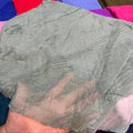 A hand holding a sheer gray fabric scarf or shawl with small metallic bead embellishments, set against a colorful background of other folded fabrics.