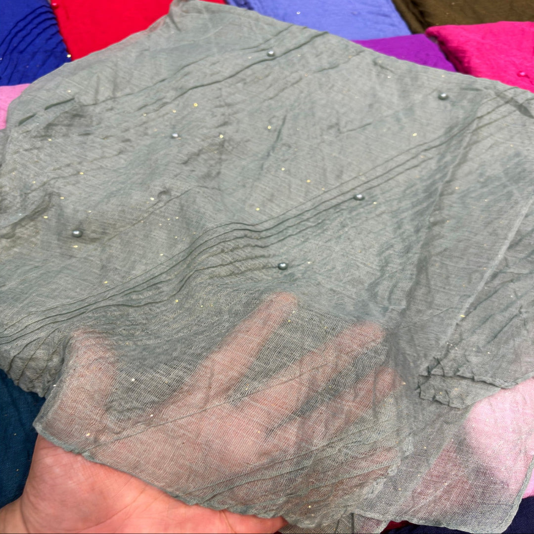 A hand holding a sheer gray fabric scarf or shawl with small metallic bead embellishments, set against a colorful background of other folded fabrics.