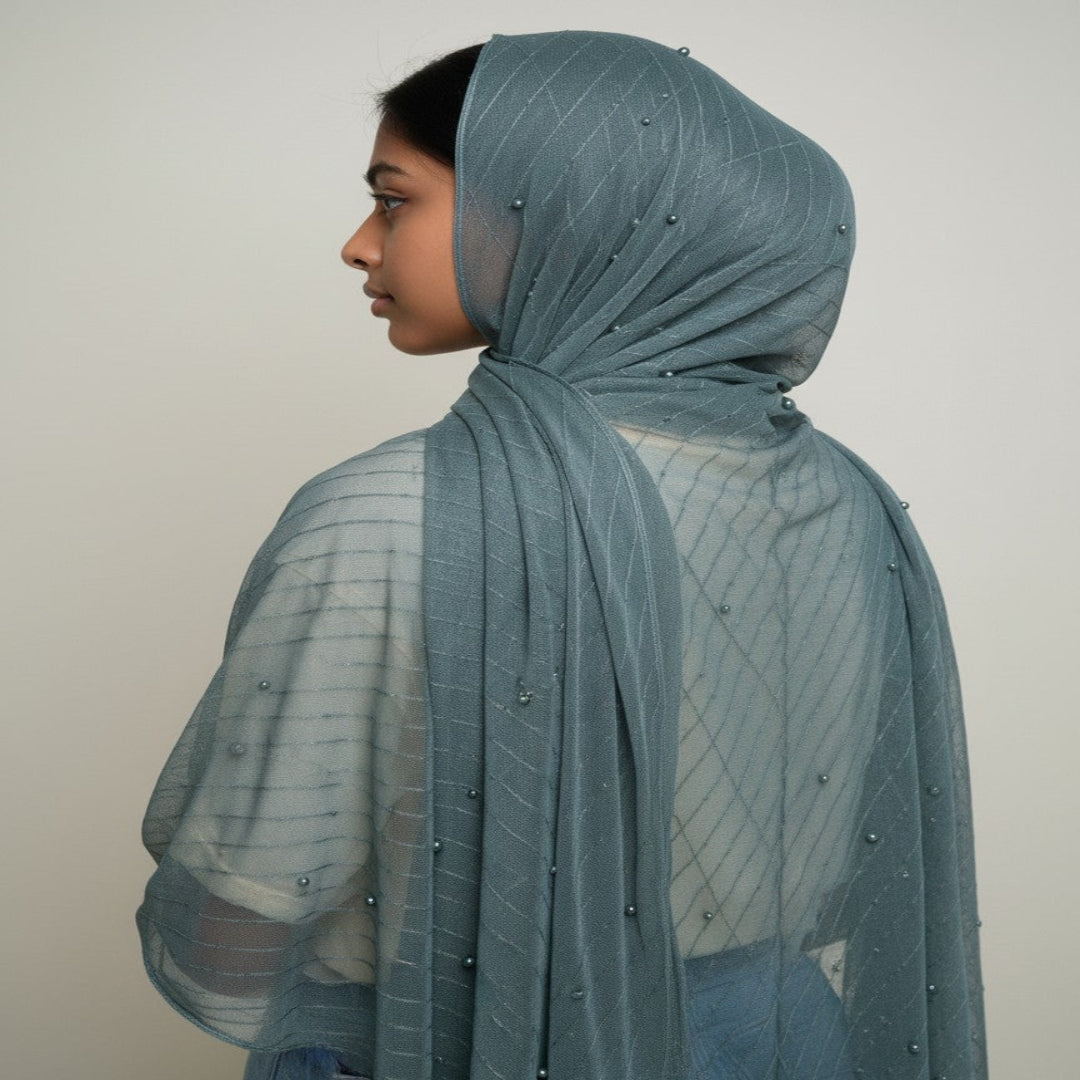 A side-profile view of a person wearing a dusty blue-gray sheer mesh hijab with small bead embellishments, paired with a white top and blue jeans, against a plain light gray background.