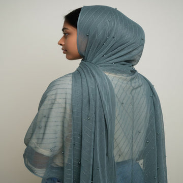 A side-profile view of a person wearing a dusty blue-gray sheer mesh hijab with small bead embellishments, paired with a white top and blue jeans, against a plain light gray background.