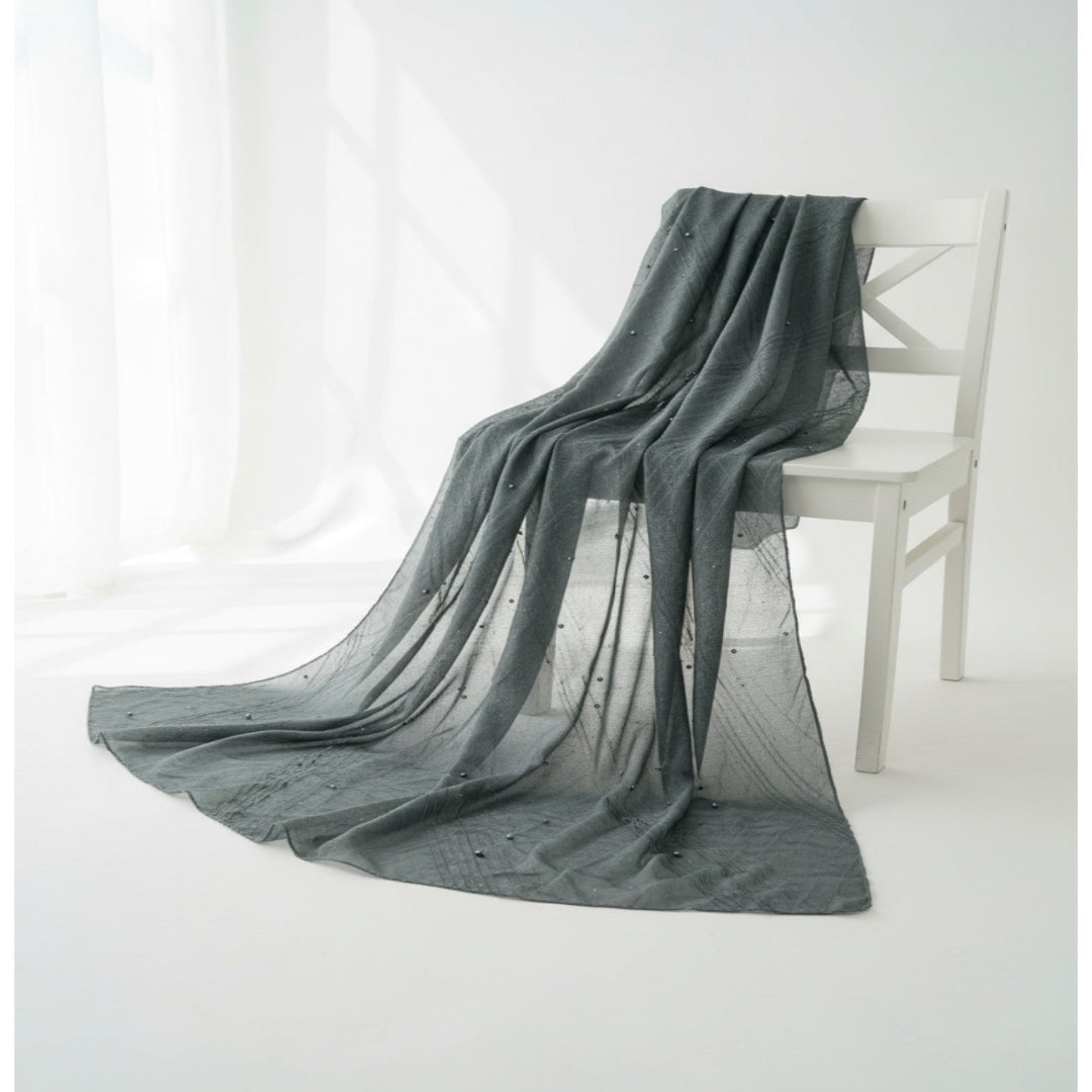 A long, sheer gray scarf or shawl with small embroidered details, draped over a white wooden chair in a bright, minimalist room.