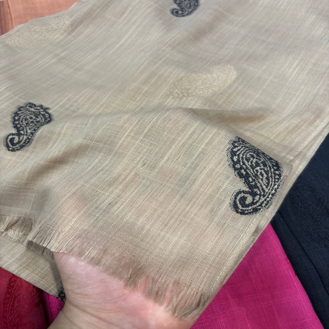 A close-up of a beige woven fabric with black paisley motifs and a fringed edge, held by a hand over a pink and dark blue fabric background.
