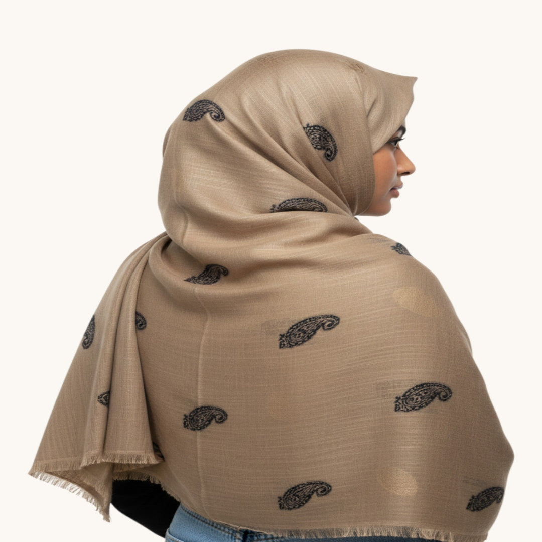 A woman shown in profile wearing a beige hijab with a subtle sheen and small black paisley motifs printed throughout, against a plain off-white background.