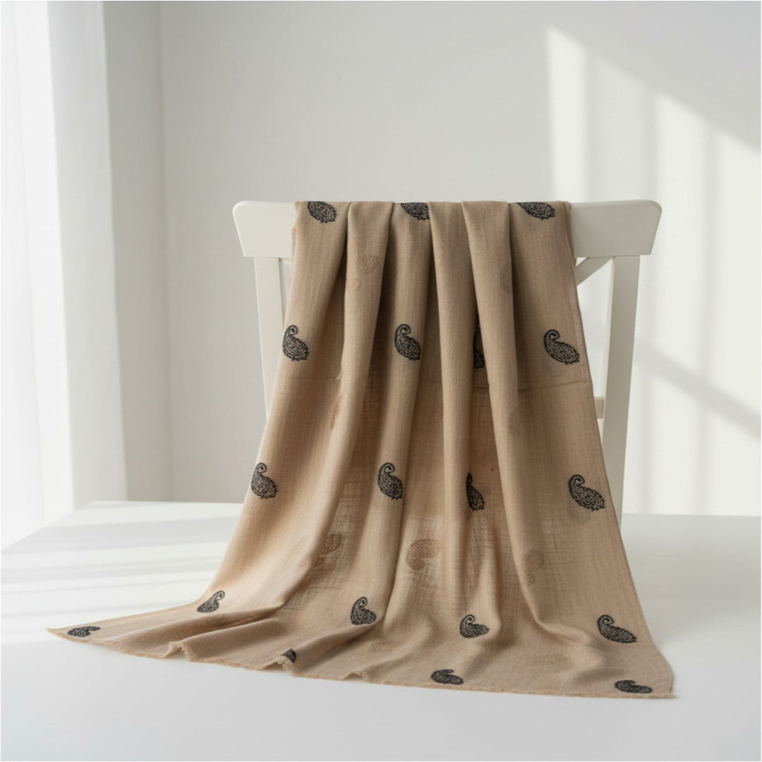 A beige draped scarf with a repeating black paisley pattern, displayed on a white wooden chair in a bright, minimalist room.
