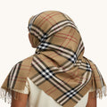 A beige plaid scarf with fringe, styled on a model from the back. The scarf features a classic check pattern in beige, black, white, and red, and is draped over the head and shoulders of a person wearing a cream-colored top against a plain off-white background.