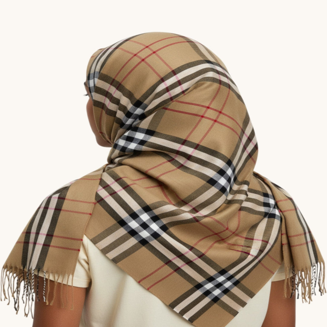 A beige plaid scarf with fringe, styled on a model from the back. The scarf features a classic check pattern in beige, black, white, and red, and is draped over the head and shoulders of a person wearing a cream-colored top against a plain off-white background.