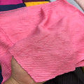 A close-up of a textured pink fabric, likely a muslin cloth or cotton gauze, being held by a hand. The fabric has a crinkled, ribbed texture and is shown in a folded state.