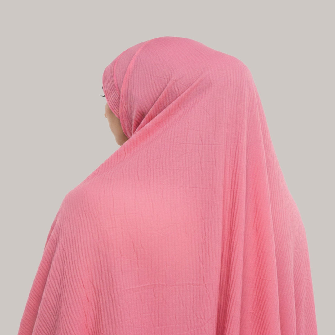 A pink textured crinkle gauze hijab worn by a model, shown from a side-back angle against a solid light gray background.