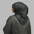 A woman shown from the back wearing a dark gray textured hijab and matching draped shawl against a solid gray background.