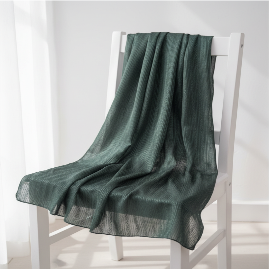 A dark green textured throw blanket draped over a white wooden chair in a bright, minimalist room.