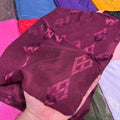 A hand holding a piece of deep burgundy fabric with a repeating tonal geometric pattern, with other colorful fabrics visible in the background.