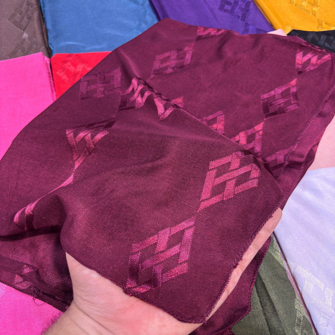 A hand holding a piece of deep burgundy fabric with a repeating tonal geometric pattern, with other colorful fabrics visible in the background.