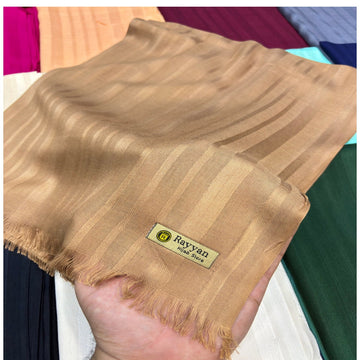 A hand holding a beige Rayyan Silk scarf with subtle vertical stripes and fringed edges, with other colorful scarves visible in the background.