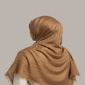 A woman wearing a shimmering taupe-brown hijab, shown from the back against a solid gray studio background.
