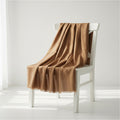 A draped camel brown throw blanket with a fine textured weave, elegantly arranged on a white wooden chair in a bright, minimalist room.
