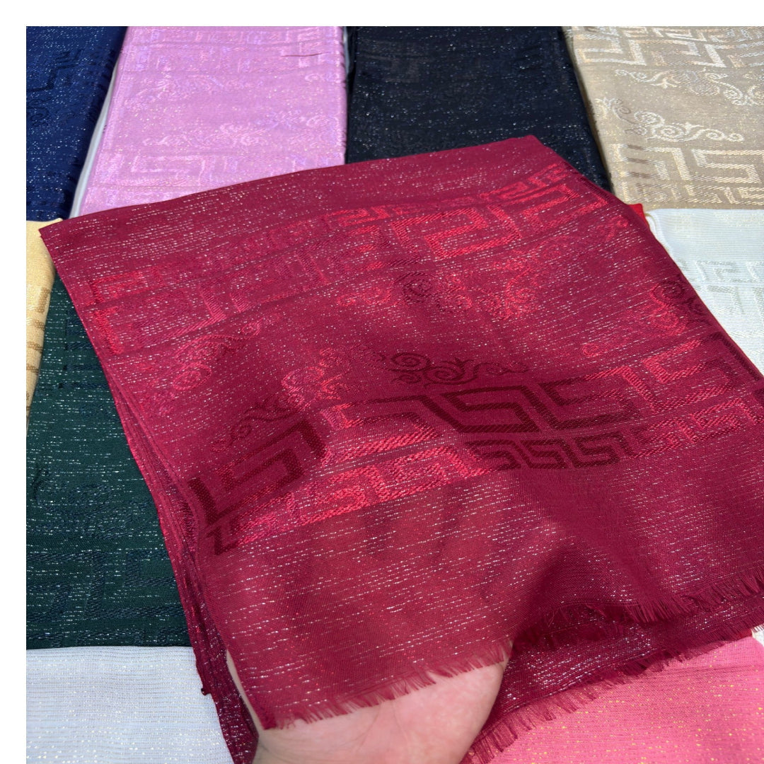 A close-up of a hand holding a folded deep red woven scarf with a subtle geometric pattern, with other color options of the scarf visible in the background.