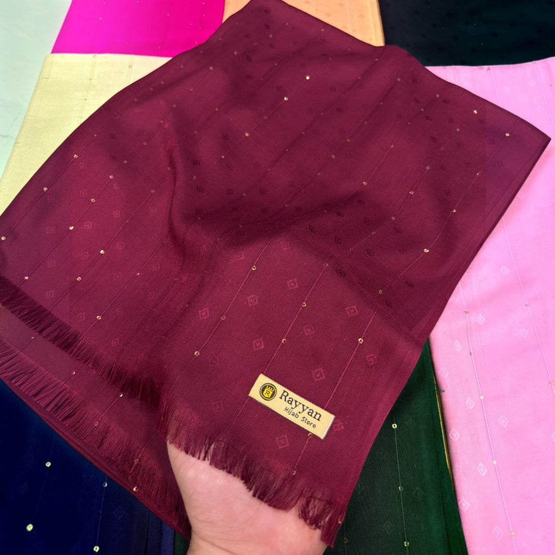 A woman shown from the back wearing a deep burgundy hijab with subtle silver glitter accents, set against a plain light gray background.