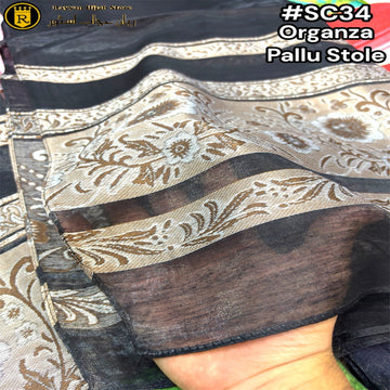A close-up of a sheer black organza pallu stole with metallic gold and silver floral patterns, held by a hand to show its drape and texture.