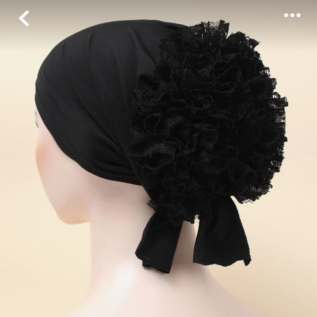A woman shown in profile wearing a black fabric head covering with a large ruffled pom-pom and side bow, photographed against a neutral gray studio background.