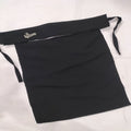A black fabric apron laid flat on a white background, featuring a wide waist strap with a small gold embroidered logo and side ties.