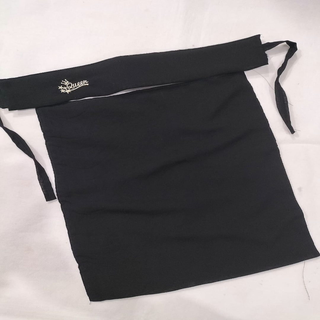 A black fabric apron laid flat on a white background, featuring a wide waist strap with a small gold embroidered logo and side ties.