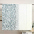 A light blue and white patterned curtain panel hanging on a rod, with a pleated white fabric panel beside it. The setting includes a wall, a small bookshelf, and a vase with flowers.