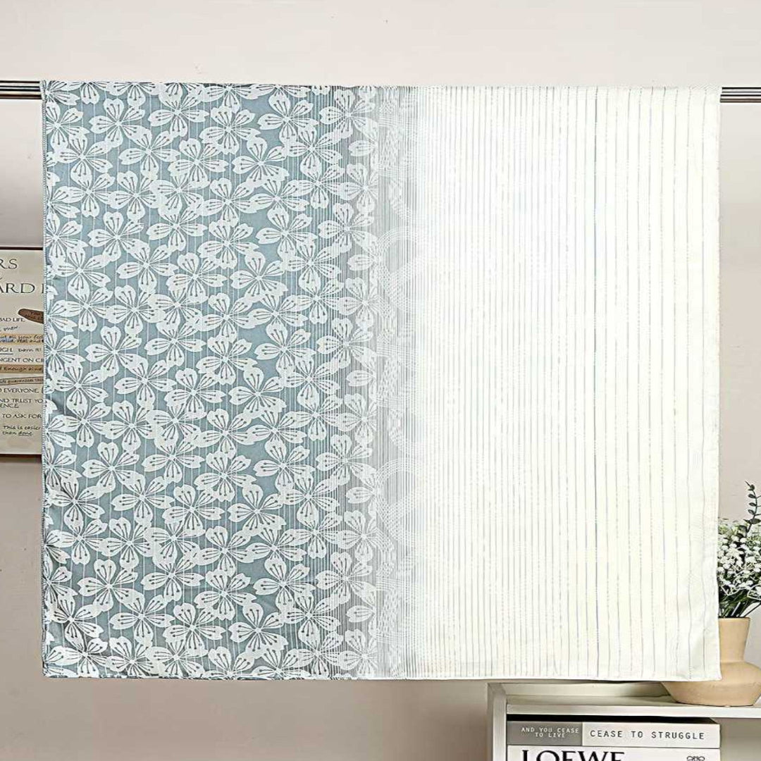 A light blue and white patterned curtain panel hanging on a rod, with a pleated white fabric panel beside it. The setting includes a wall, a small bookshelf, and a vase with flowers.